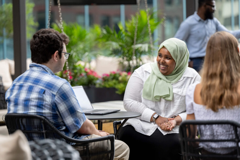 Turnberry professionals, a young Caucasian man and an African American woman wearing a head covering, converse in Turnberry's outdoor meeting space amongst additional multi ethnic colleagues