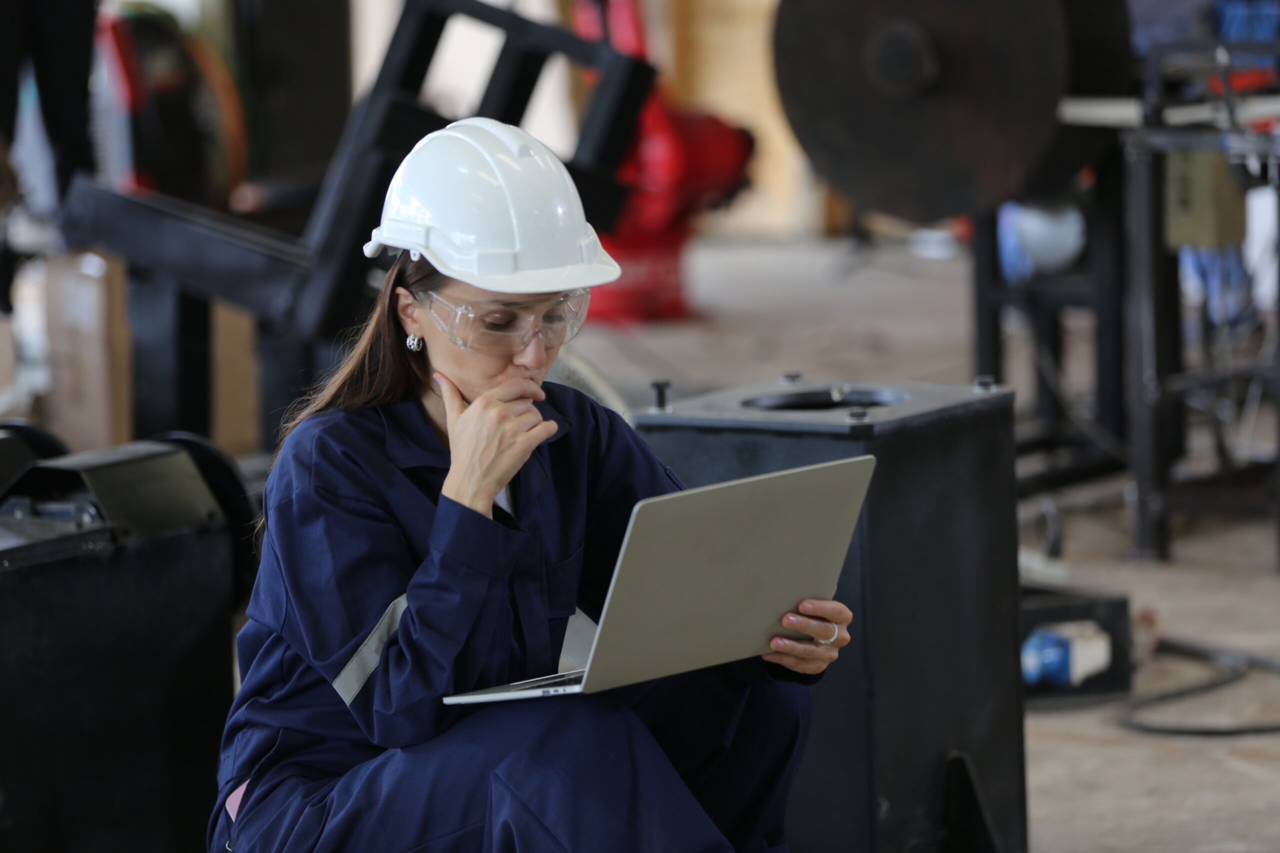 Female manufacturing professional wearing a hardhat and coveralls reviews content on a laptop in a manufacturing environment