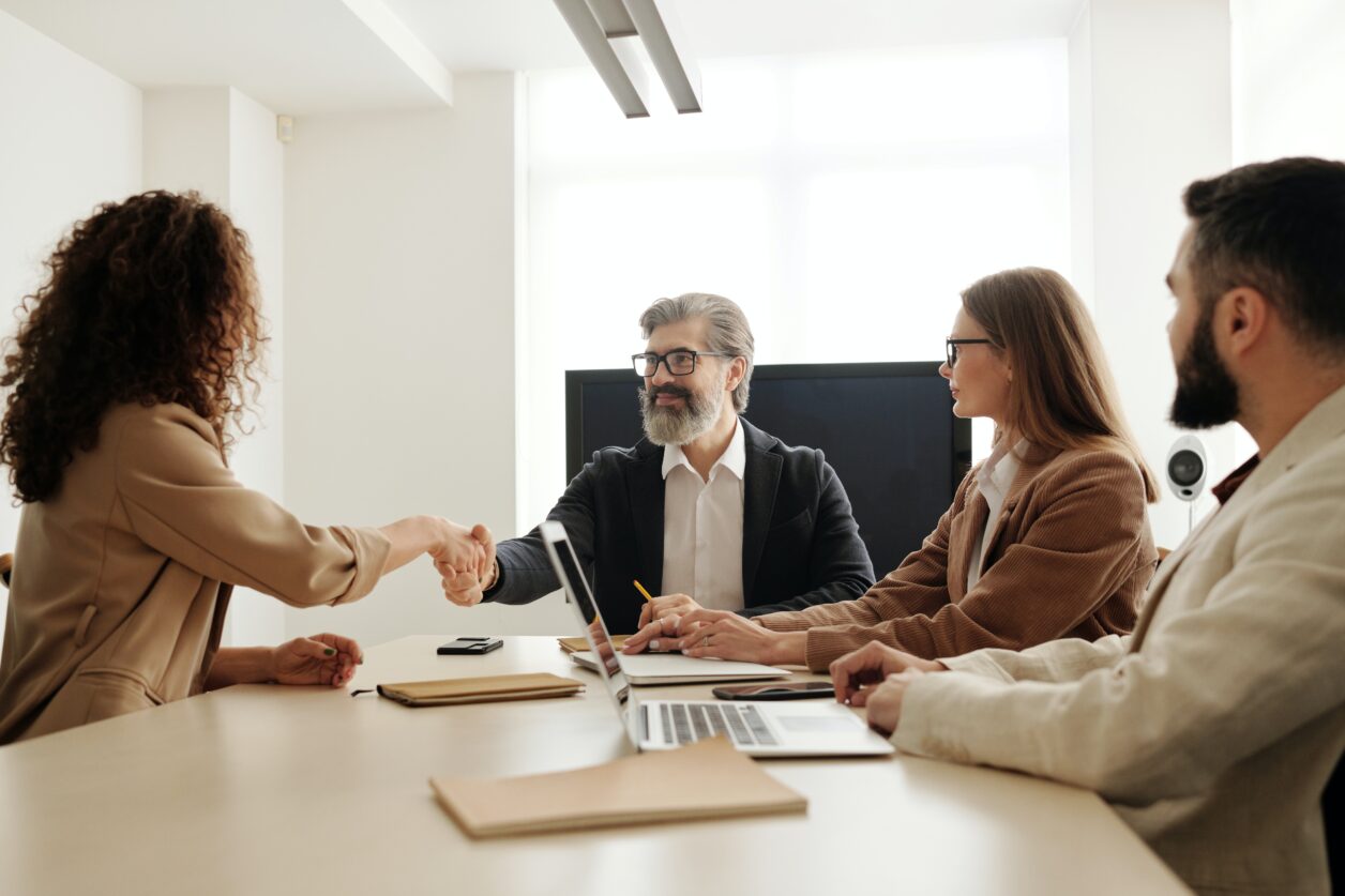Mature male professional shakes hand of female professional in conference room with two other professionals using laptops