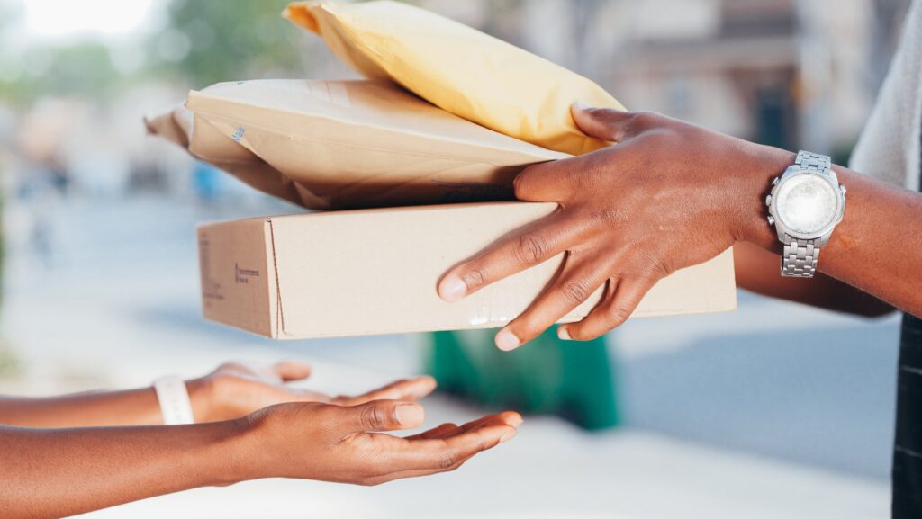 An African American delivery person wearing a silver watch hands off a box and two thick envelopes to an another African American person
