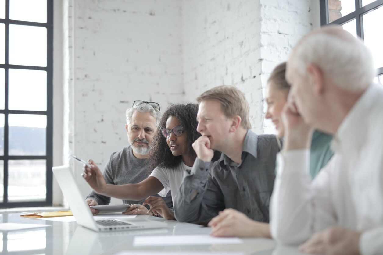 Seated group of mixed age, gender, race professionals listen to young African American female professional consulting a laptop during discussion