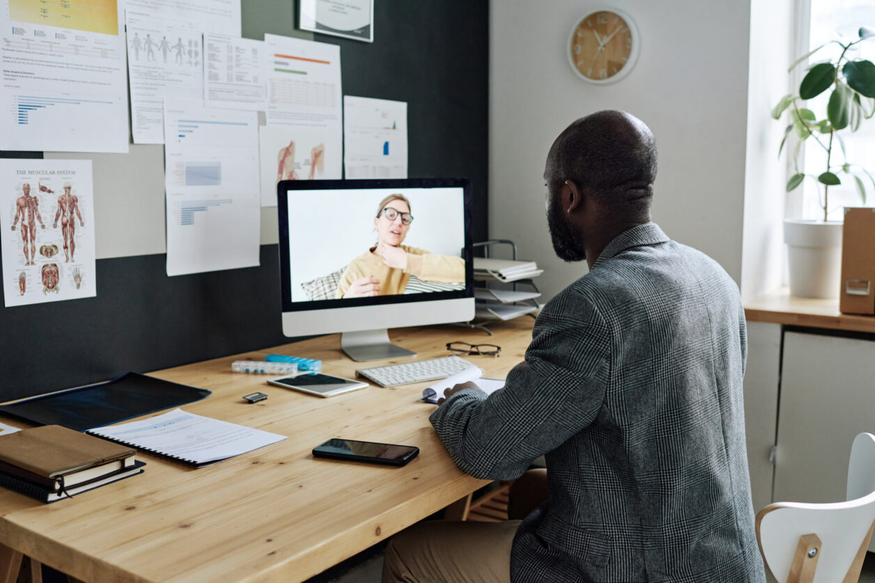 An African American man sits at a modern desk while on a video call with a Caucasian woman demonstrating vocal or neck exercises