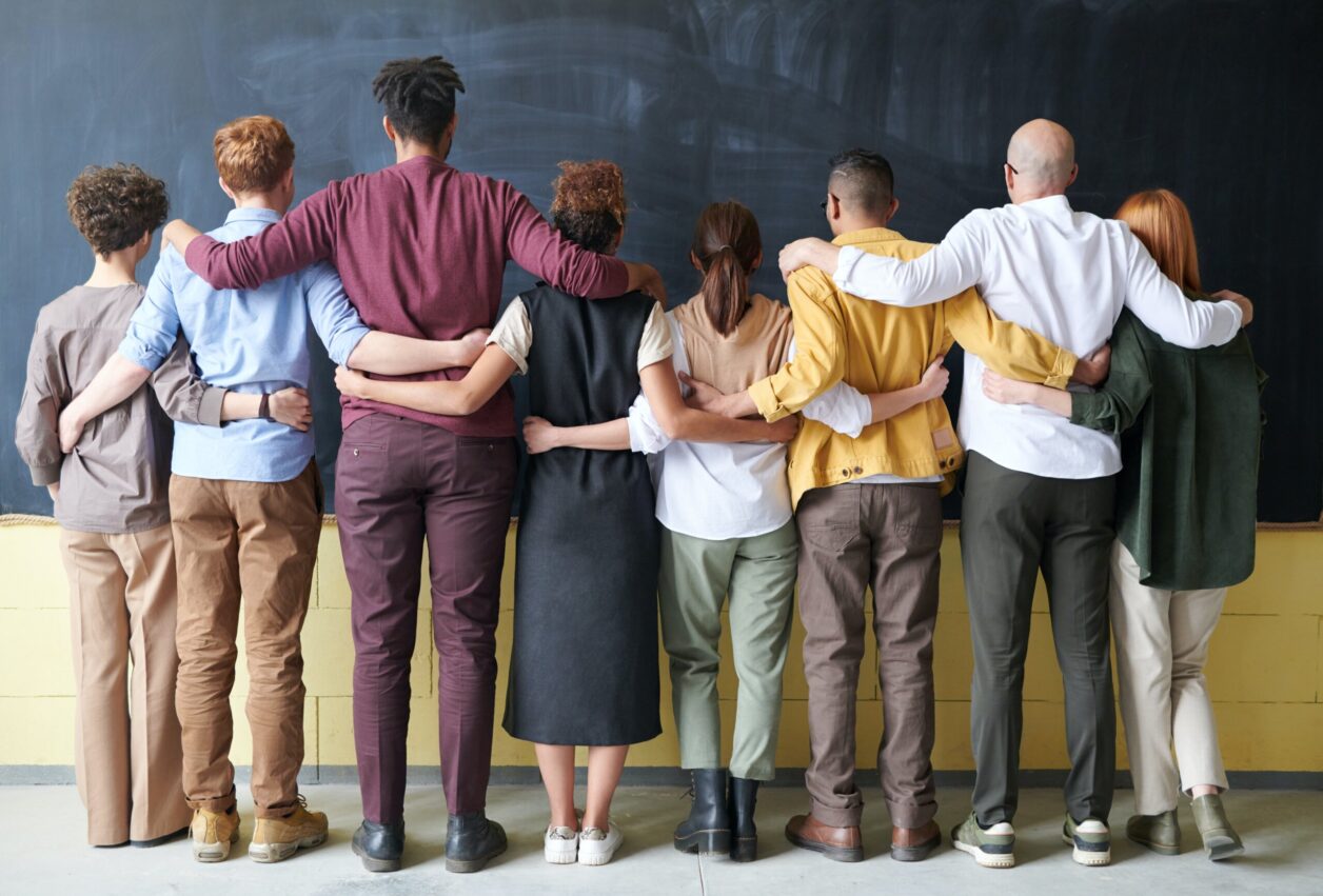 Stock image of a diverse group of people forming a line in front of a chalkboard with their hands across one another's backs or shoulders in support of one another