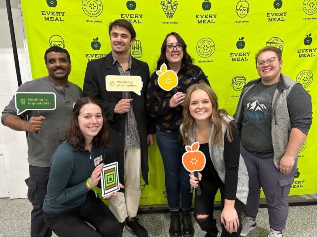Six Turnberry employees pose for a photo at an Every Meal event to fight child hunger, participants hold speech buble signs containing positive statements or graphical icons of fruit