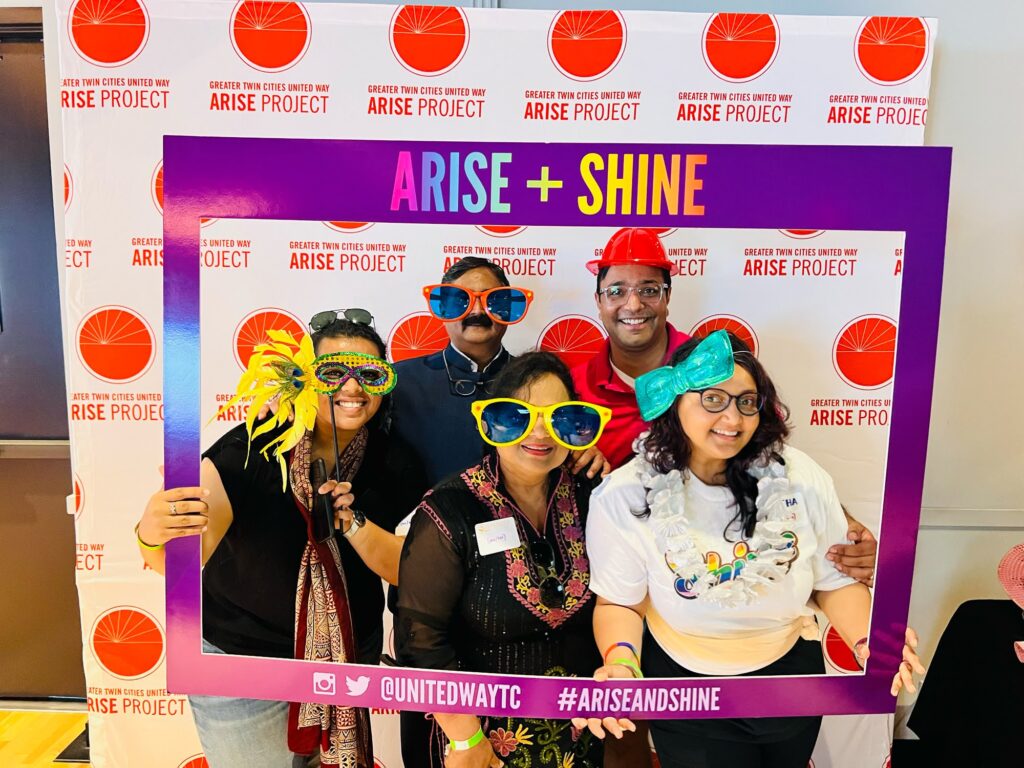 Turnberry professionals pose for fun photo with novelty paper frame at Arise event as part of Shine Crew LGBTQ support in collaboration with the Twin Cities United Way, attendees wear oversized sunglasses, hair bow, a mardi-gras style mask and plastic novelty hard hat