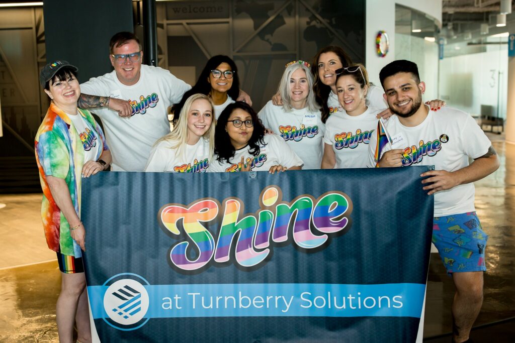 Group of Turnberry professionals wearing Shine iniative teeshirts and holding a Shine at Turnberry Solutions sign to convey support for LBGTQ employess