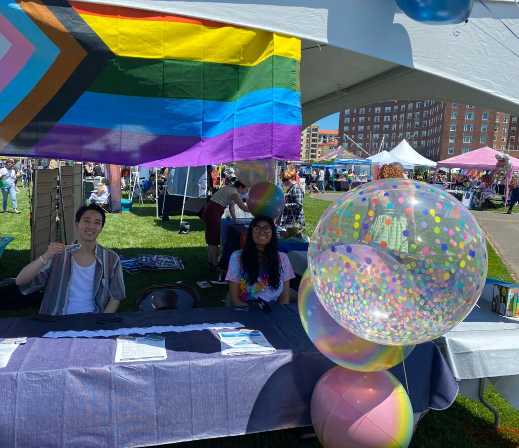 Two people at a table at a Pride event in New Jersey with multi-color baloons and a LGBTQ+ flag over the display