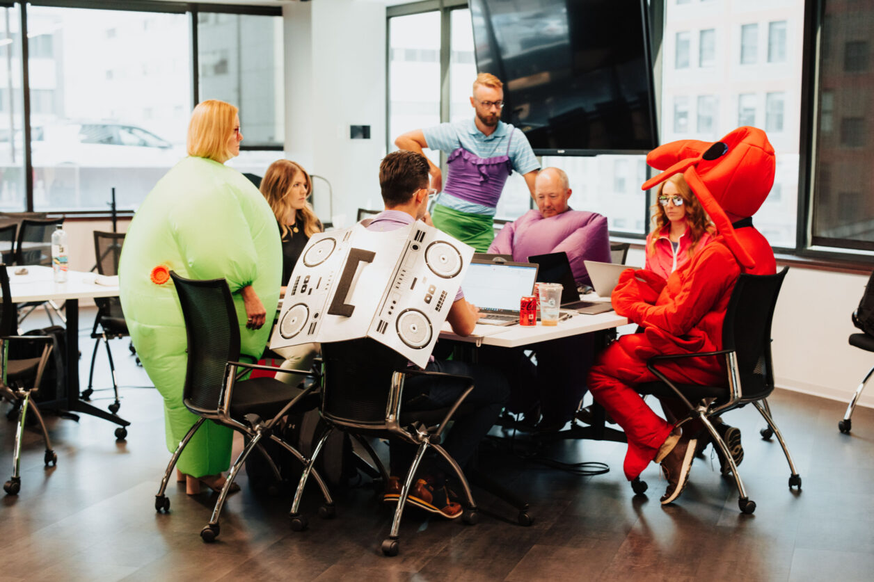Group of mid-career Turnberry professionals work around a table while wearing costumes including a beat box radio, red dog, blow up suits in purple and pale green, and a man wearing a purple form fitting ruffled top over a blue polo shirt with a green skirt or shorts in Minneapolis