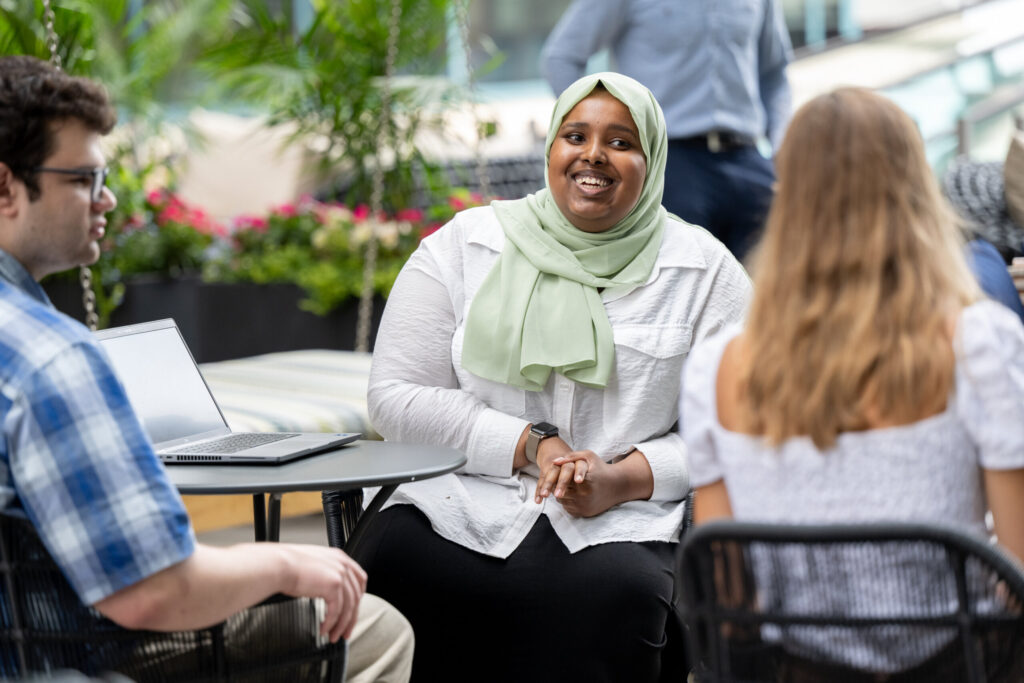 Turnberry professionals, a young Caucasian man and an African American woman wearing a head covering, converse in Turnberry's outdoor meeting space amongst additional multi ethnic colleagues