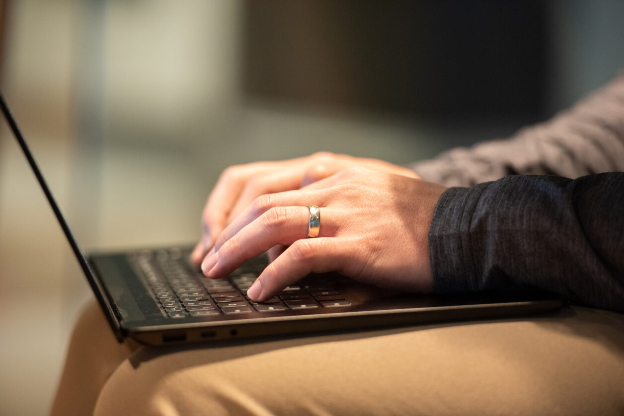 A man's hands wearing a ring types on a keyboard resting on his lap