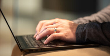 A man's hands wearing a ring types on a keyboard resting on his lap