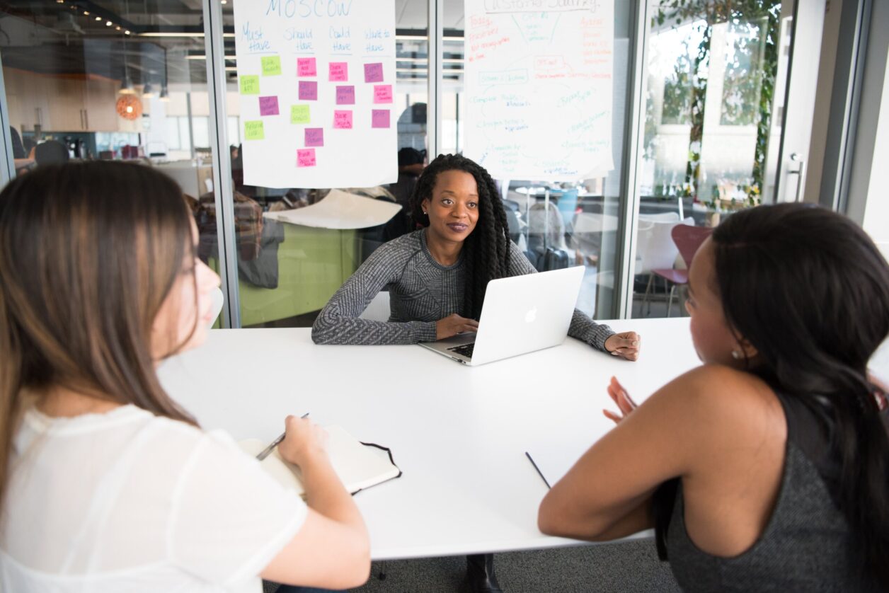 An African American woman sits behind a laptop listening during a conversation to women sitting across from her in a modern conference room