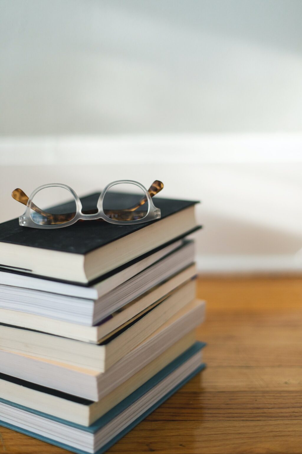 A stack of books sits on a wood table with a pair of clear acetate and tortoise shell glasses sits on top of the books