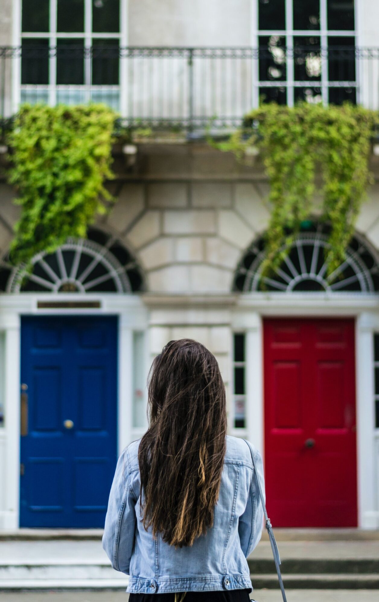 A woman in a denim jacket stands across the street from white stone rowhouses with a blue door and a red door with balconies above that have greenery
