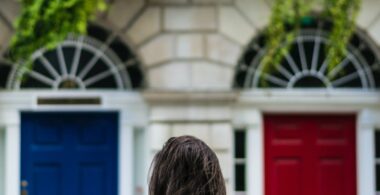 A woman in a denim jacket stands across the street from white stone rowhouses with a blue door and a red door with balconies above that have greenery